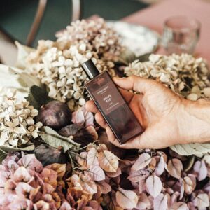 Hand holding a perfume bottle amidst an elegant floral arrangement on a table.