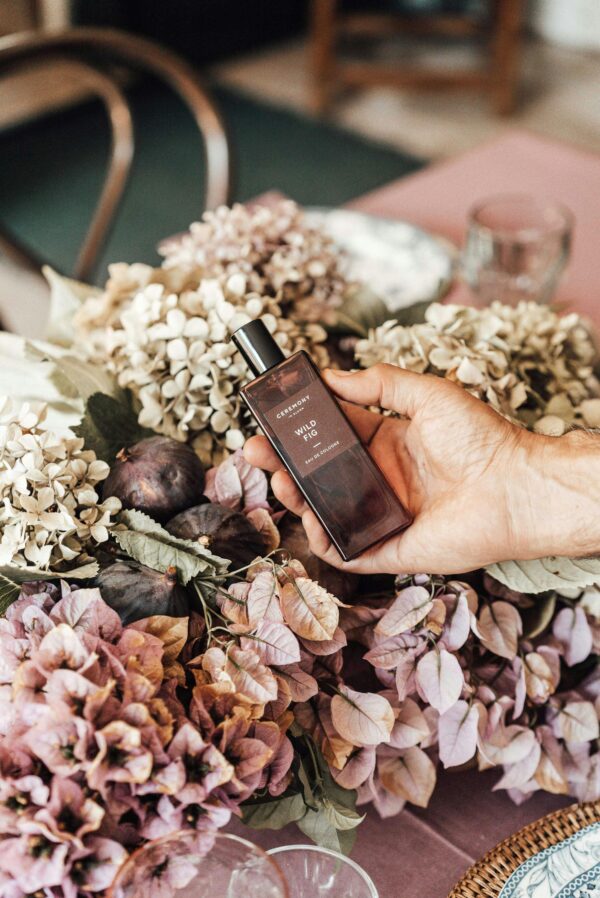 Hand holding a perfume bottle amidst an elegant floral arrangement on a table.